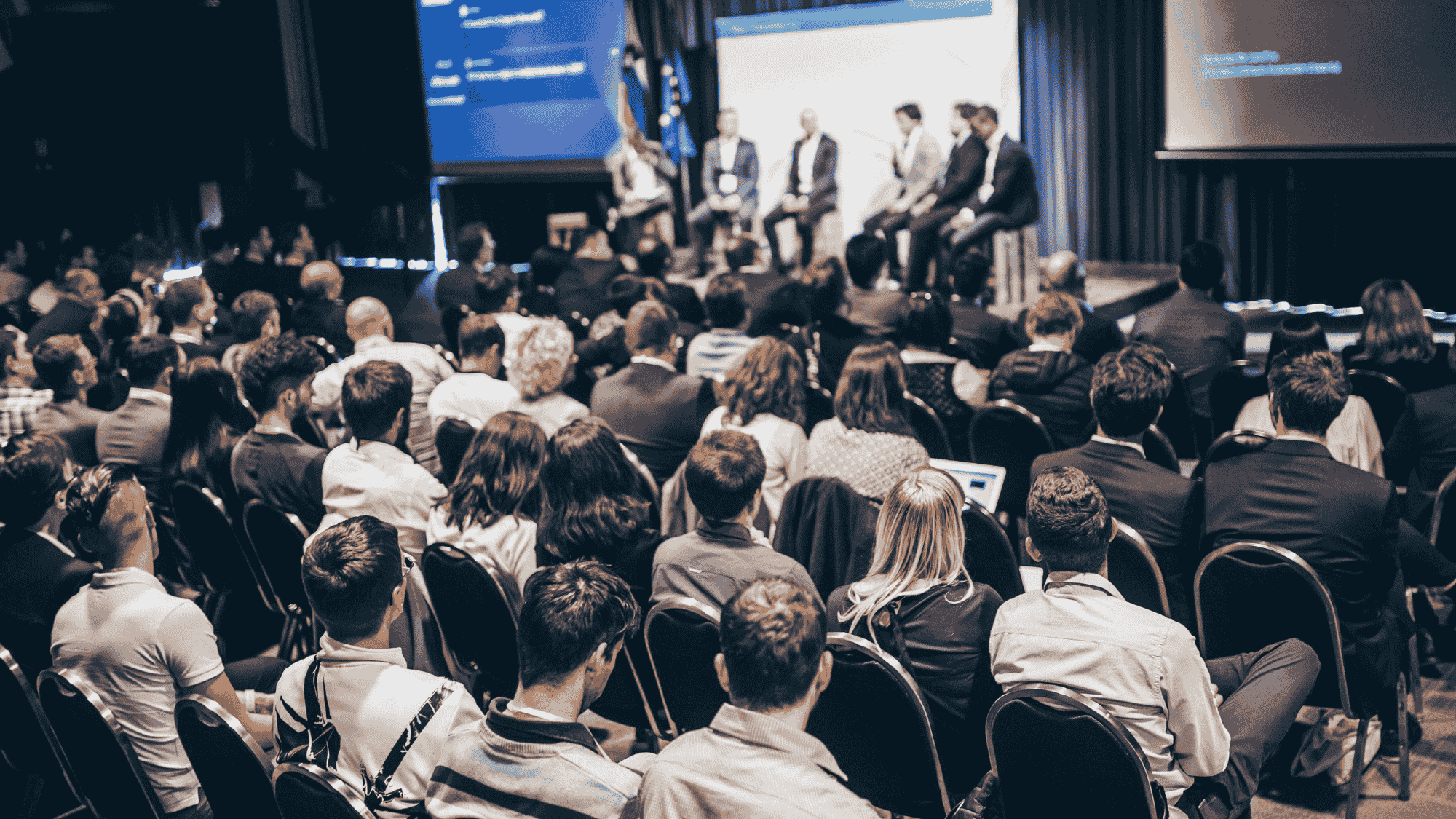 An audience at a corporate event listen to speakers on stage.