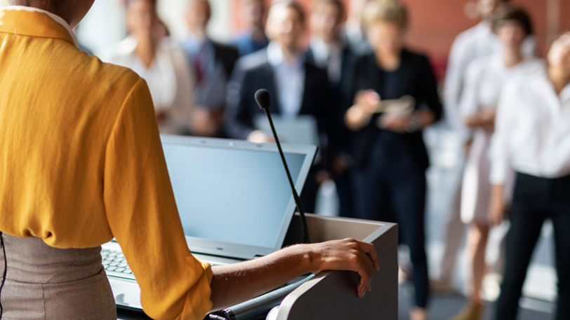 A speaker at a podium with a microphone at a corporate conference.