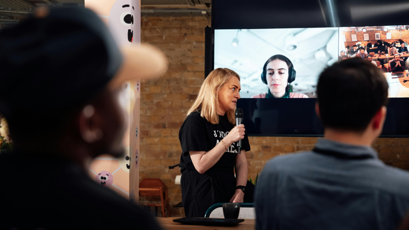 A presenter holds a microphone in front of a screen showing remote attendees.